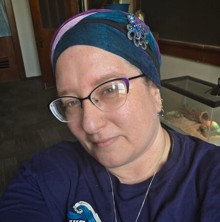 An adult white person facing the camera with a smile, wearing glasses, a dark blue shirt, and a matching headwrap with an octopus pin for decoration.